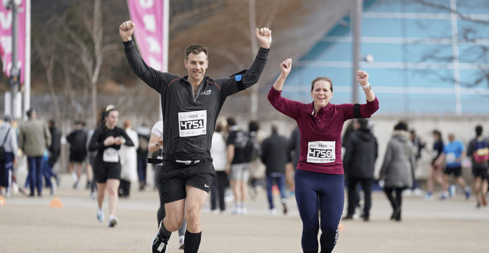 Two smiling runners with race bibs raise their arms in celebration as they approach the finish line during a marathon event. Other participants and spectators are seen in the background.