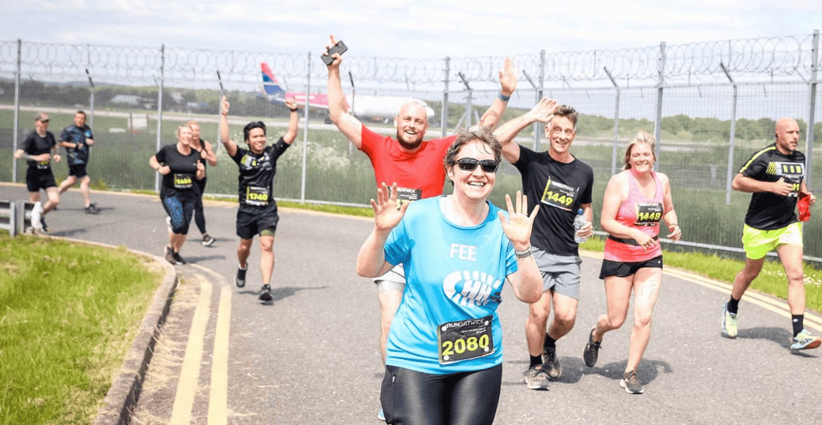 A group of runners, some smiling and waving, participate in a race on a road next to a fence topped with barbed wire. The weather is sunny, and most runners wear race bibs and athletic clothing.