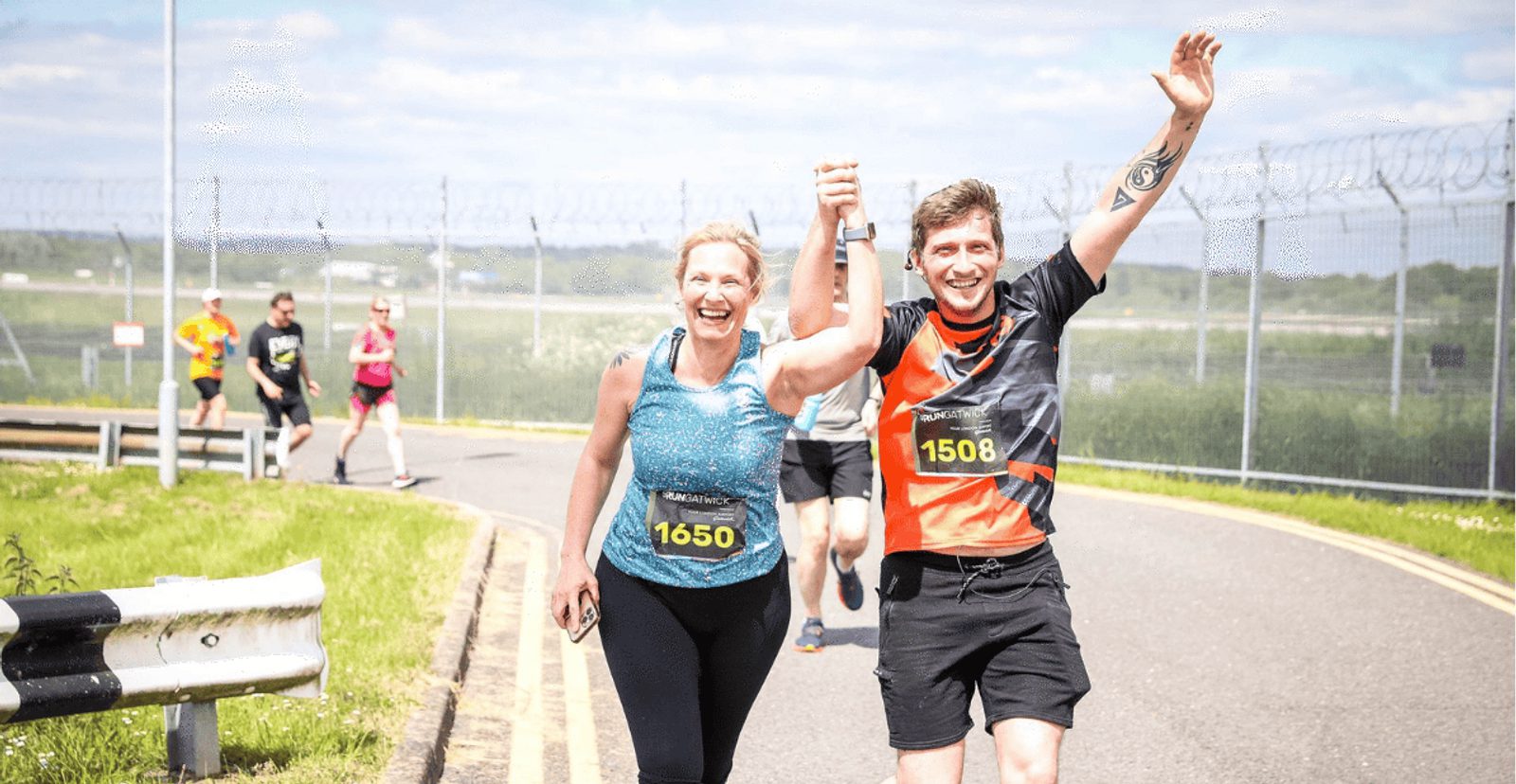 Two smiling runners, holding hands and raising them in celebration, finish a race together on a sunny day. Both wear numbered race bibs and athletic clothes; other runners are visible in the background.