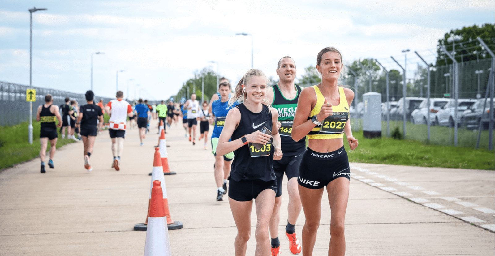 Runners participate in a road race, with several people jogging on a paved path lined with cones. Two women in front are smiling, wearing sportswear and race bibs. Other runners follow behind on a cloudy day.