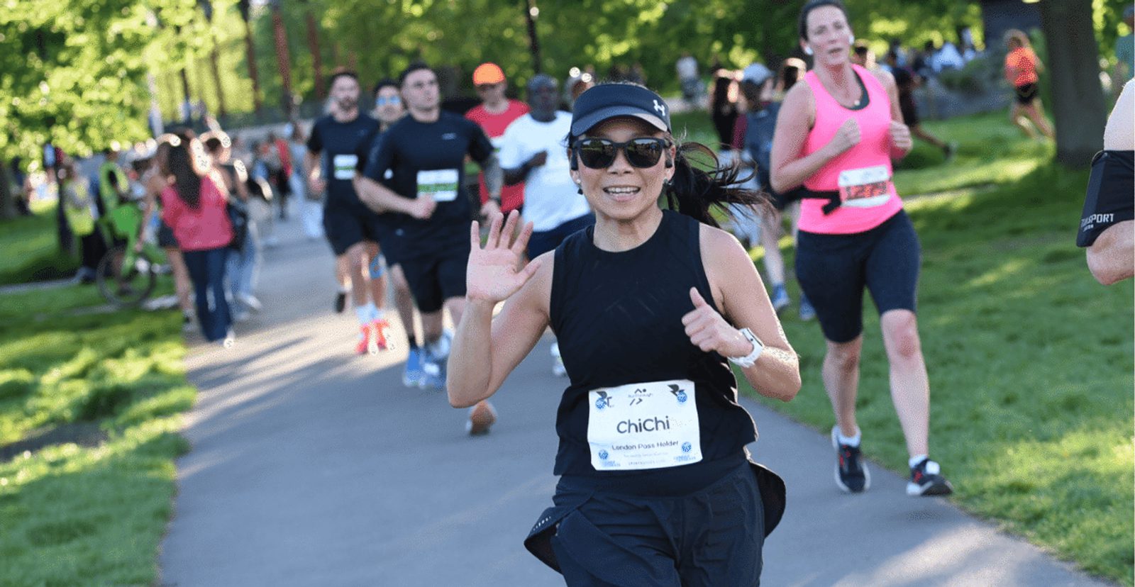 A woman in black athletic gear smiles and waves while running on a park path during a race, with other runners and green trees visible in the background.
