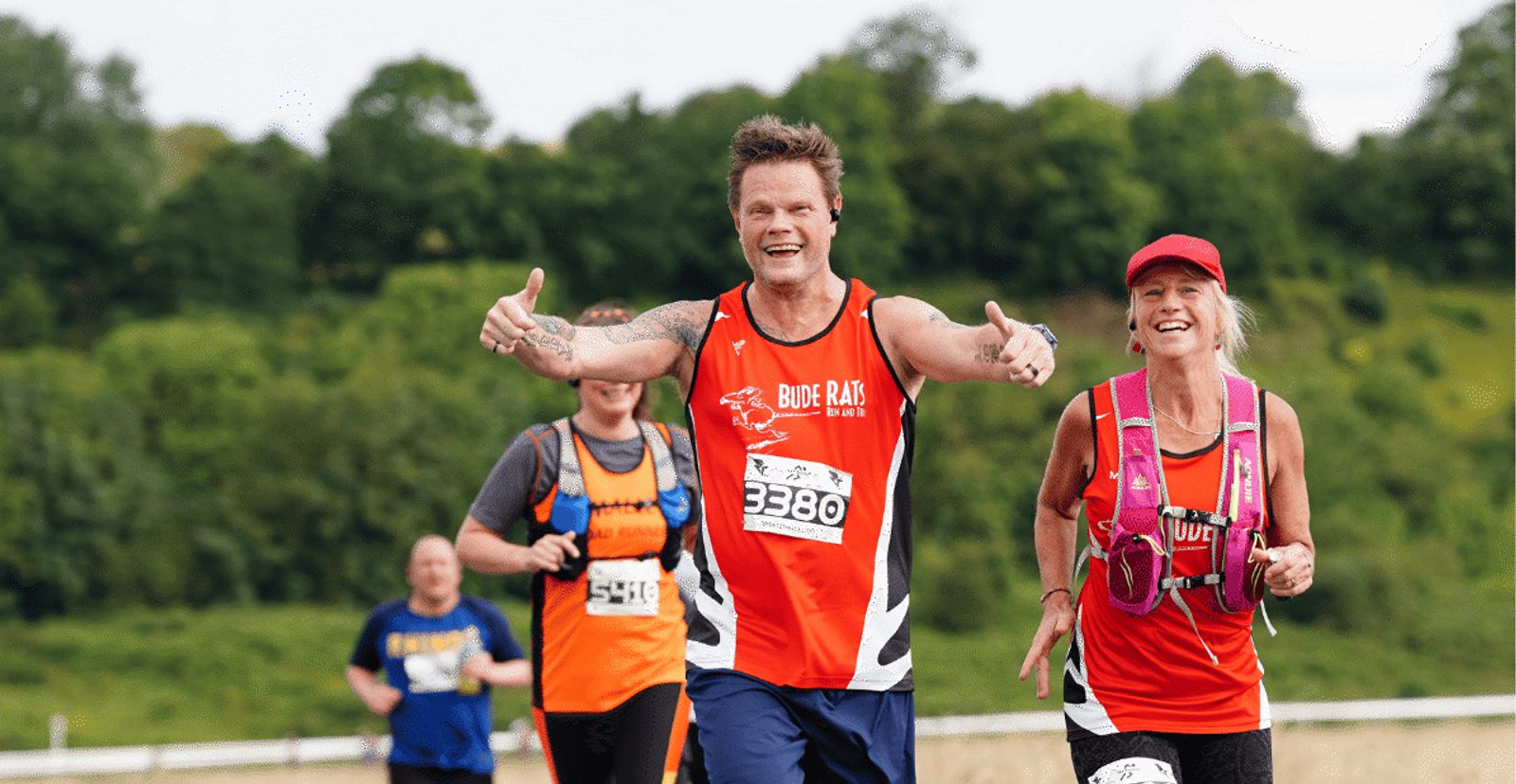 Three marathon runners in red and orange gear smile and gesture enthusiastically as they run outdoors on a sunny day, with greenery and trees in the background.