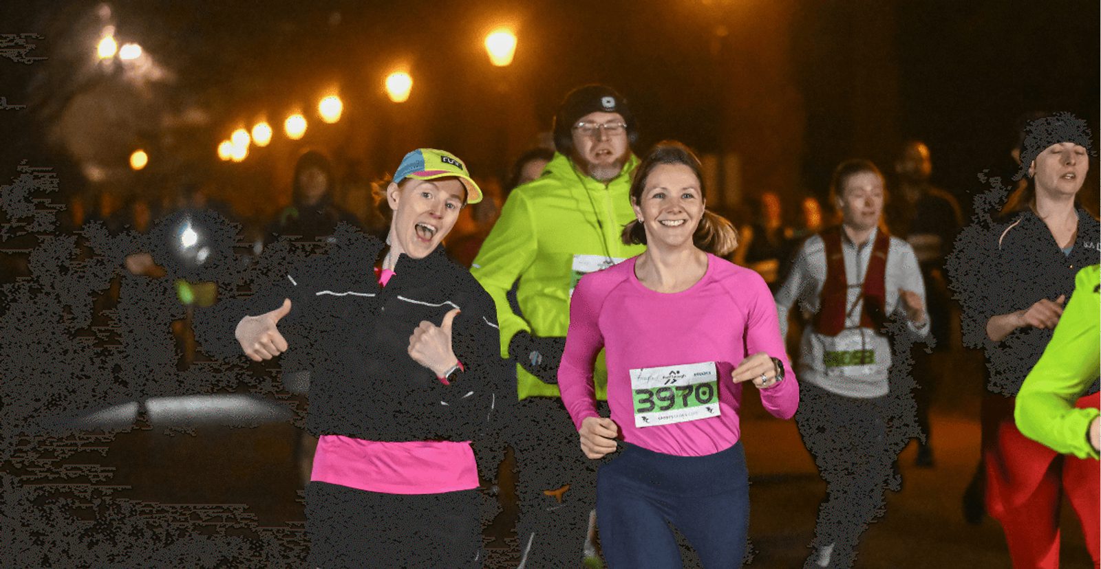A group of runners participate in a night race. Two women in front, both smiling, wear bright athletic clothing and race bibs. Streetlights illuminate the background as other runners follow behind.