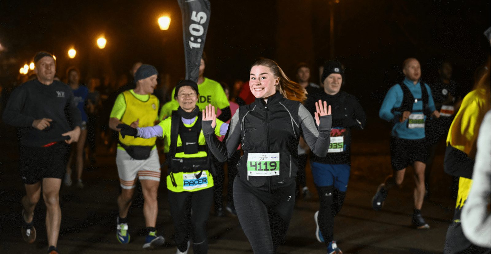 A smiling woman in black running clothes raises her hands while running a nighttime race with other runners on a well-lit street. She wears a race bib number 4119.