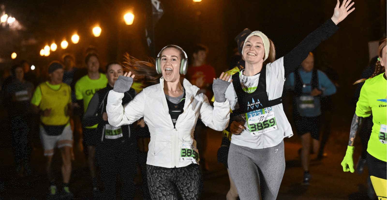 Two women in athletic wear, smiling and waving as they run in a nighttime race. Other runners follow behind them under streetlights, creating a lively and energetic atmosphere.