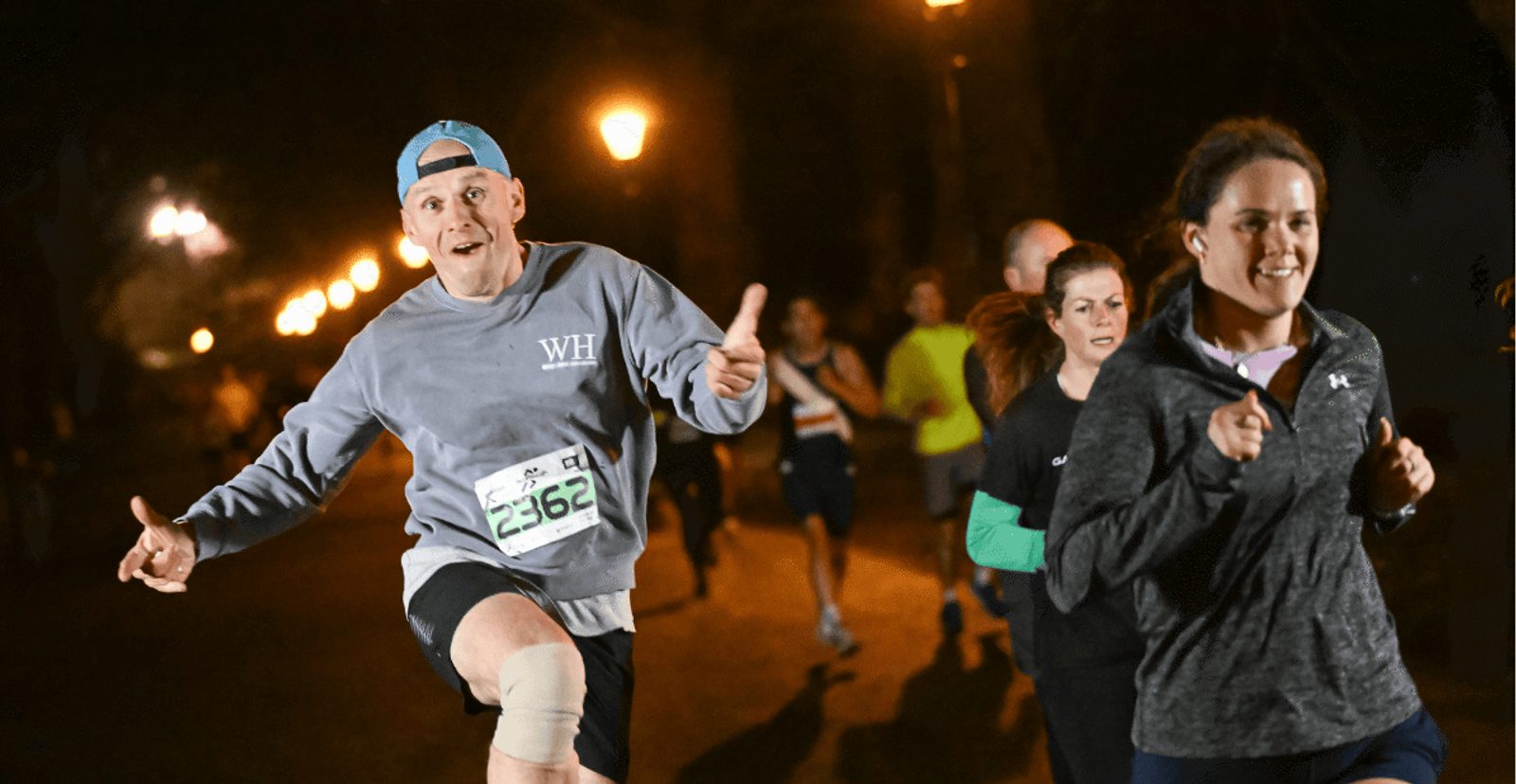A group of people run at night on a lit path. A man in front, wearing a cap backward and gray sweatshirt, makes a playful gesture and smiles, while a woman beside him jogs, focused and smiling. Other runners follow behind.