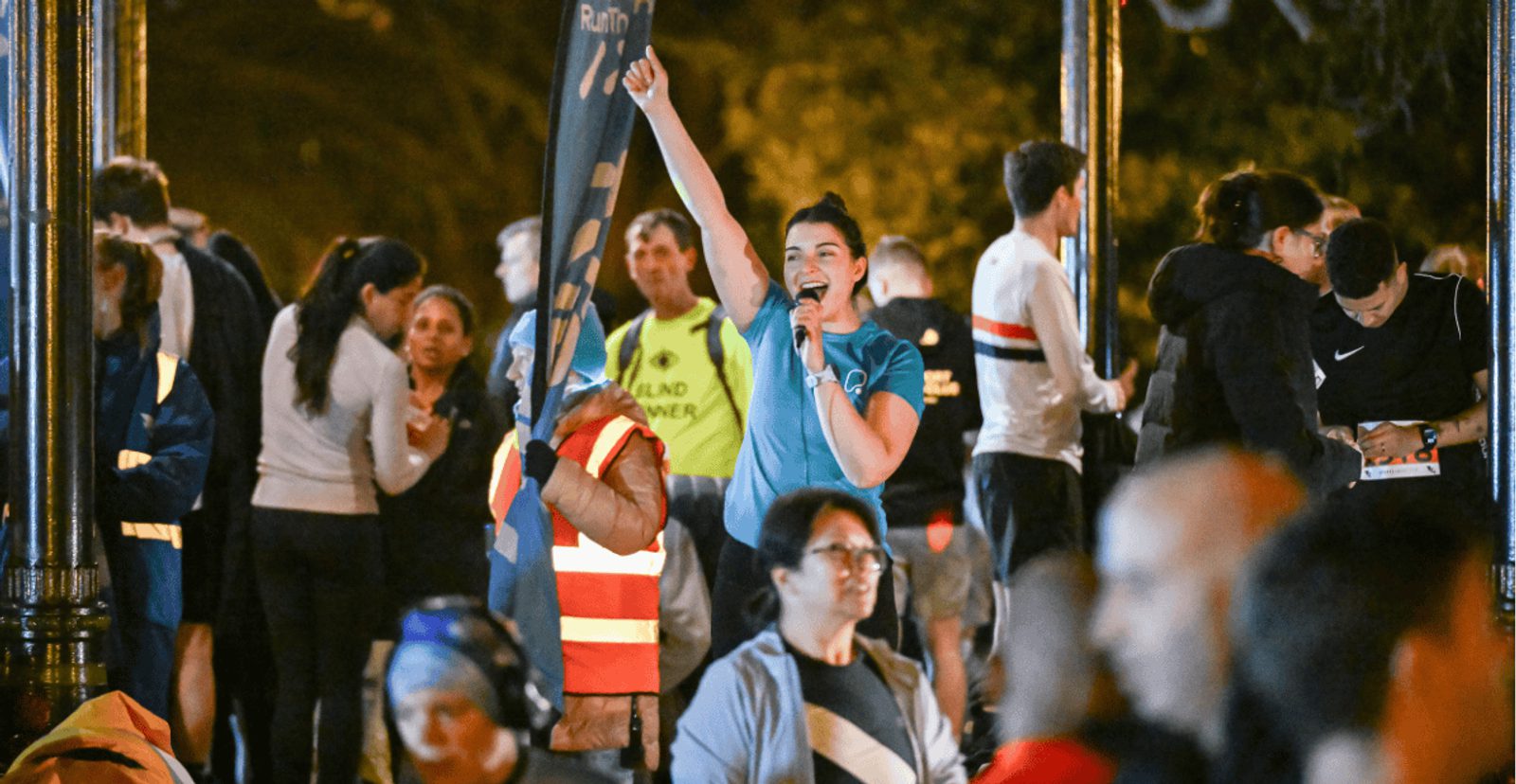 A woman wearing a teal shirt speaks into a microphone and raises her fist, standing among a crowd of people gathered outdoors at night, some in running attire and others wearing reflective vests.