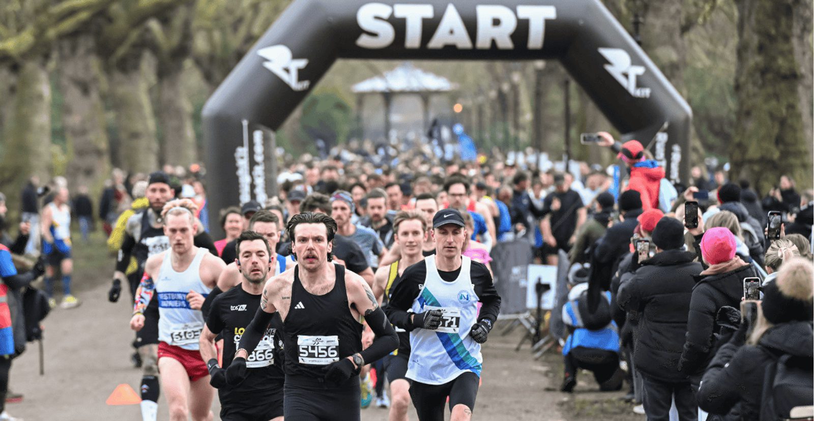 A large group of runners begin a race under a black inflatable "START" arch on a tree-lined path, surrounded by cheering spectators dressed in winter clothing.