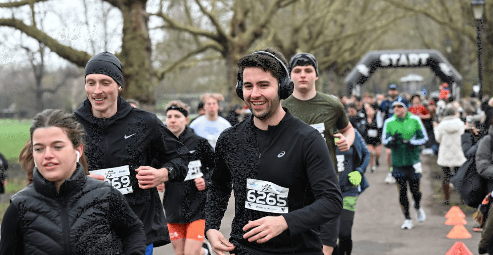 A group of runners wearing race bibs jogs along a tree-lined path during a race. Some wear headphones and cold-weather gear. An inflatable "START" arch is visible in the background.