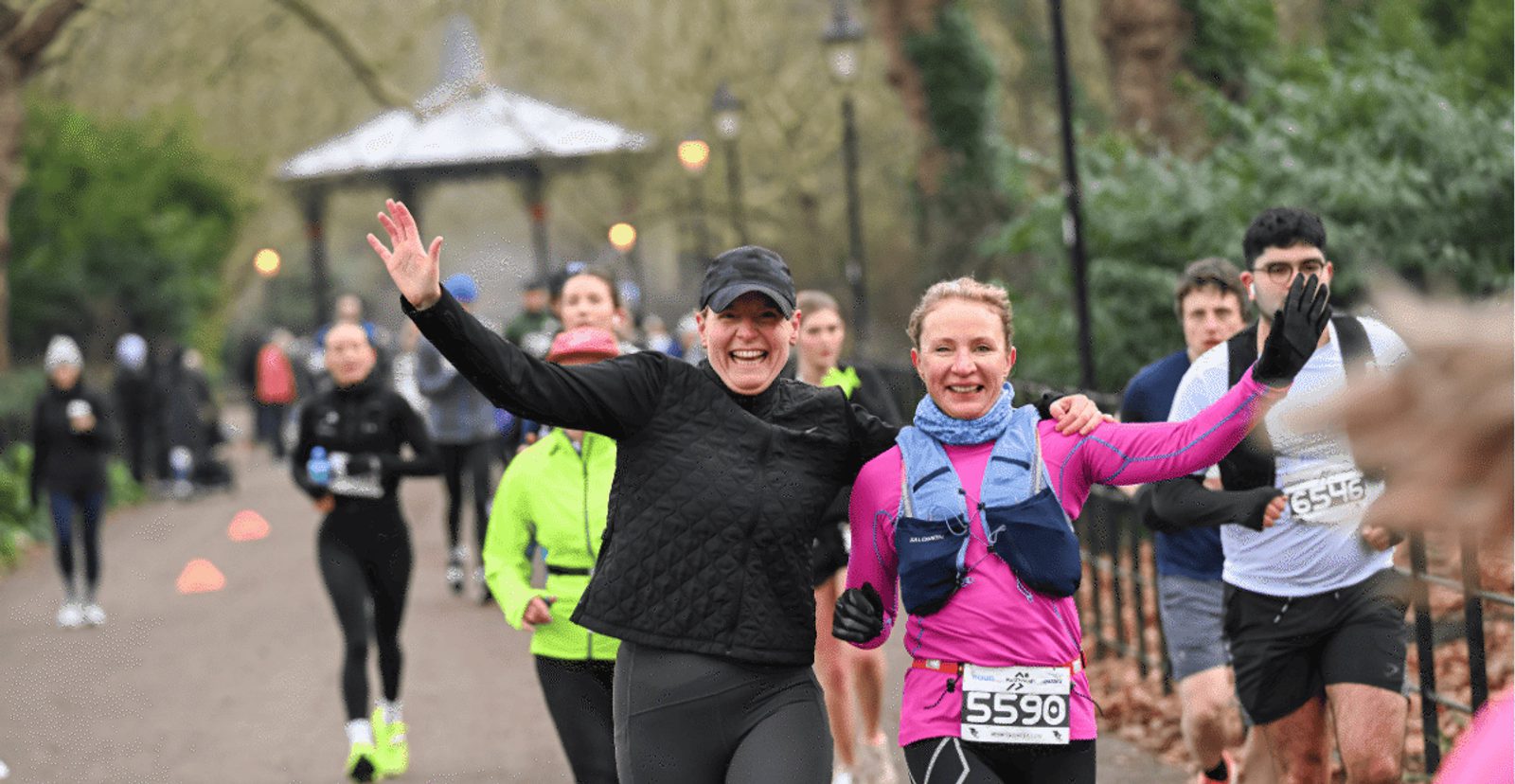 Two women smiling and waving while running in a race, surrounded by other runners on a park path. The weather appears cool, and participants wear jackets and running gear.