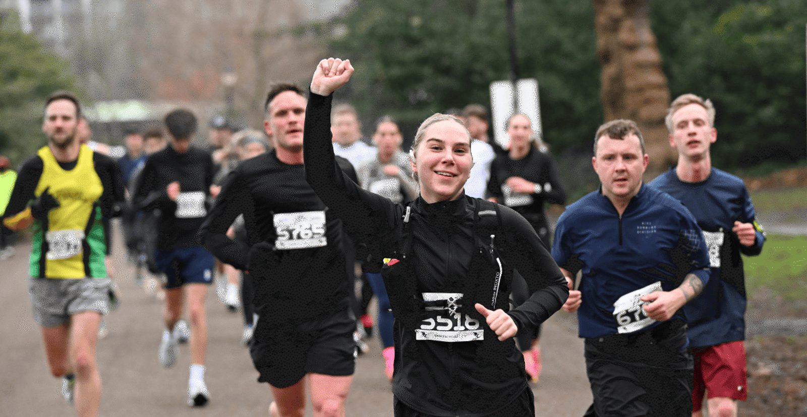 A group of runners participates in a race on a park path. A smiling woman in the foreground raises her fist in celebration while other runners follow closely behind. The weather appears cool and cloudy.