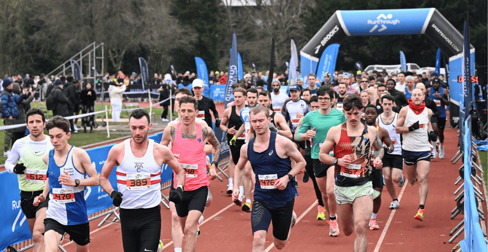 A large group of runners, wearing race bibs, start a race on a track. Spectators watch from the sidelines, and a blue inflatable arch marks the starting line. Trees and event banners are visible in the background.