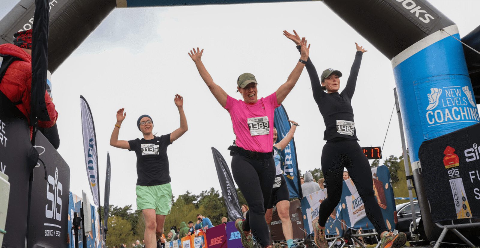 Three women runners with raised arms celebrate as they cross a race finish line under an inflatable arch, surrounded by cheering spectators and race banners.