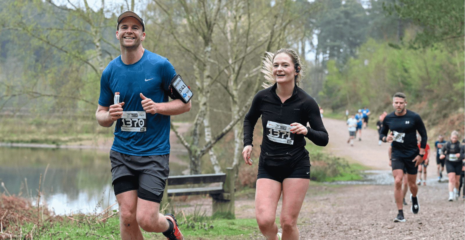 Two runners, a man in a blue shirt and a woman in black, smile as they jog on a wooded trail by a lake. Other runners are visible in the background, and the setting is natural and green.