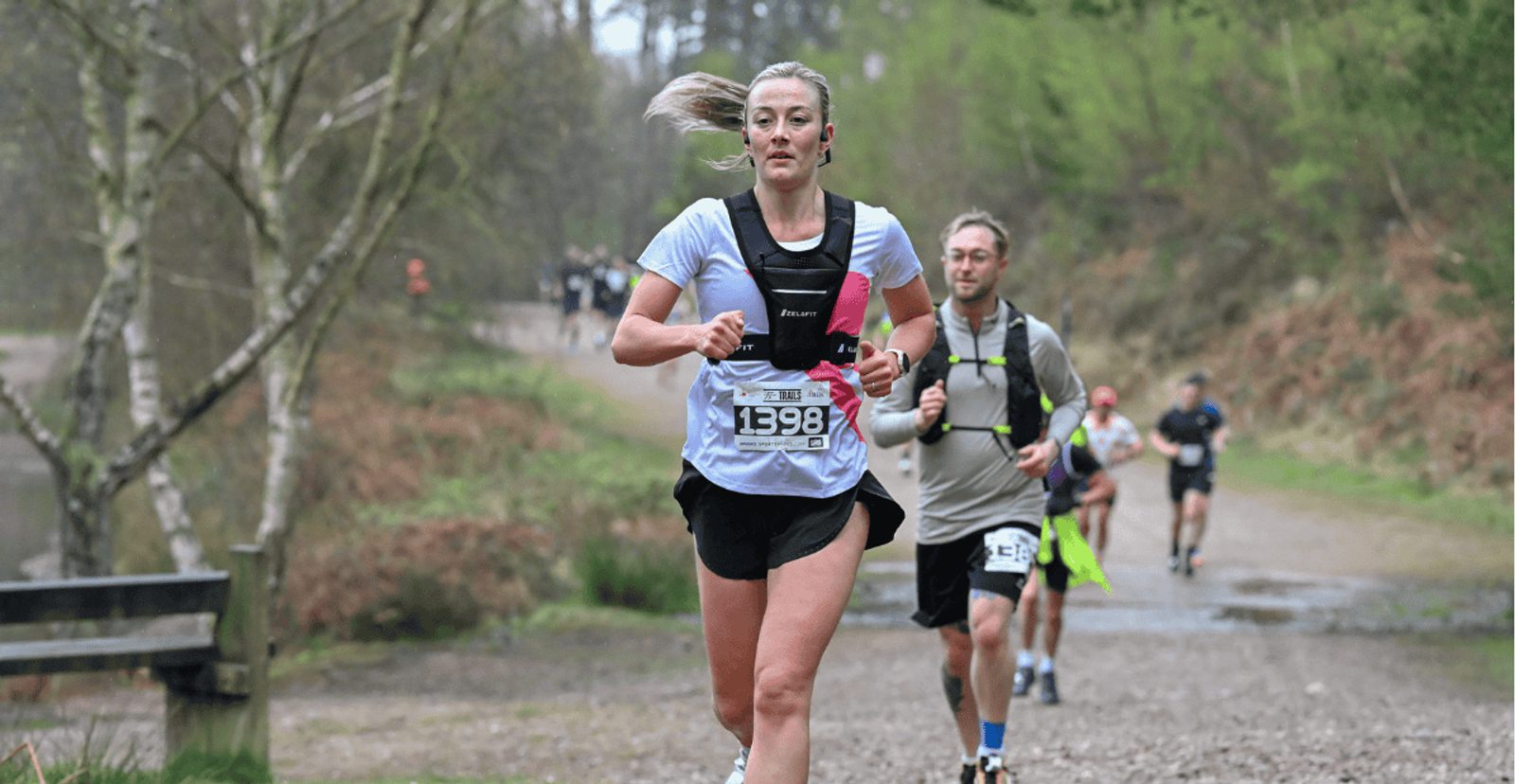 A woman running on a forest trail during a race, wearing a numbered bib and sportswear, with other runners behind her on the path. Trees and greenery line both sides of the trail.