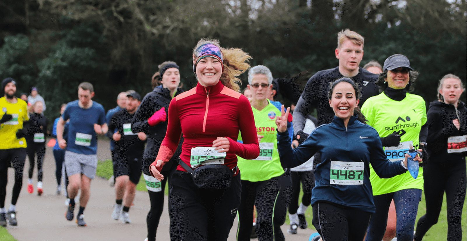 A group of runners, both men and women, participate in a race outdoors. Two women in the foreground are smiling; one wears a red jacket and headband, the other a dark blue jacket. Other runners follow behind them on a park path.