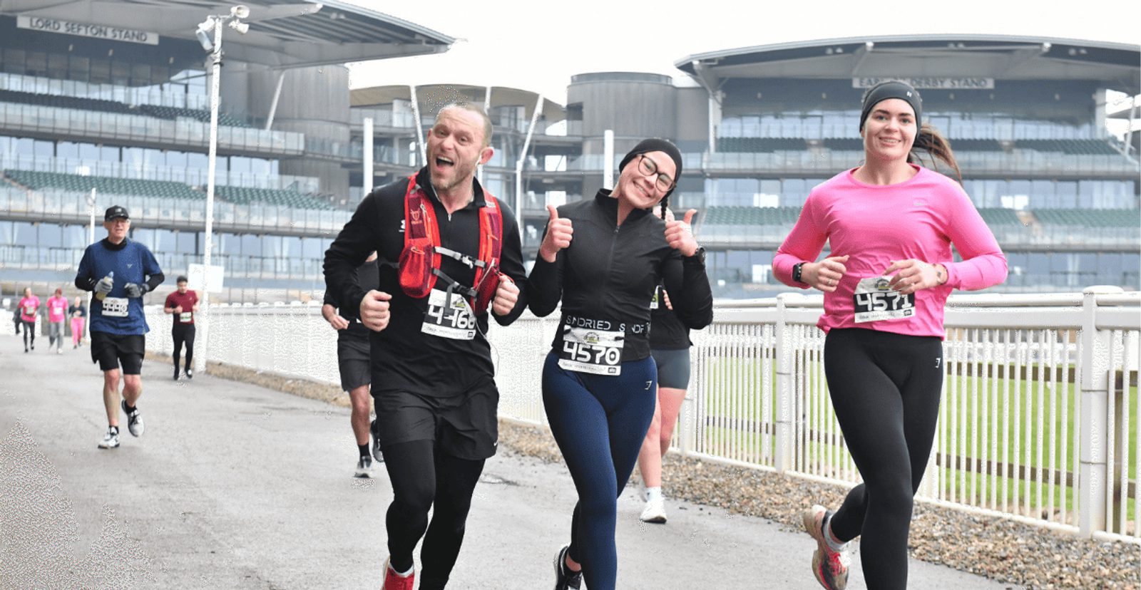 Three runners in athletic gear smile and pose for the camera during a race on a paved path, with large stadium stands visible in the background. Other runners follow behind them on the cloudy day.
