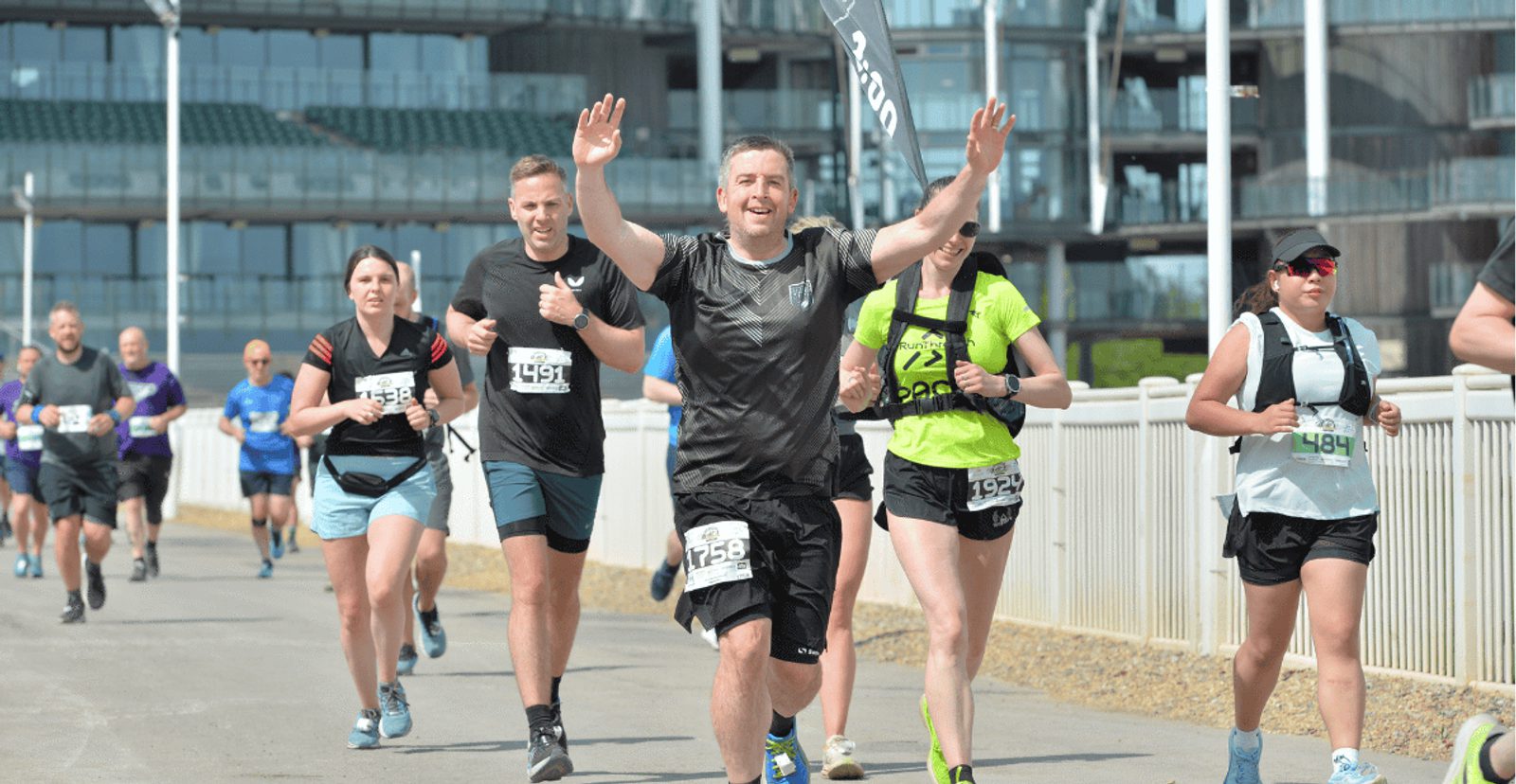 Runners participate in a road race on a sunny day. One man in front raises his arms in celebration, while others run behind him. Modern buildings and a fence line the background. Everyone wears race bibs and athletic gear.
