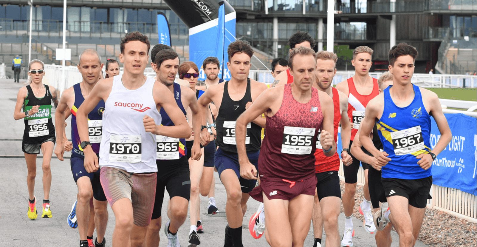 A group of male runners in athletic gear and numbered bibs begin a road race, running closely together on a paved path, with modern buildings visible in the background.