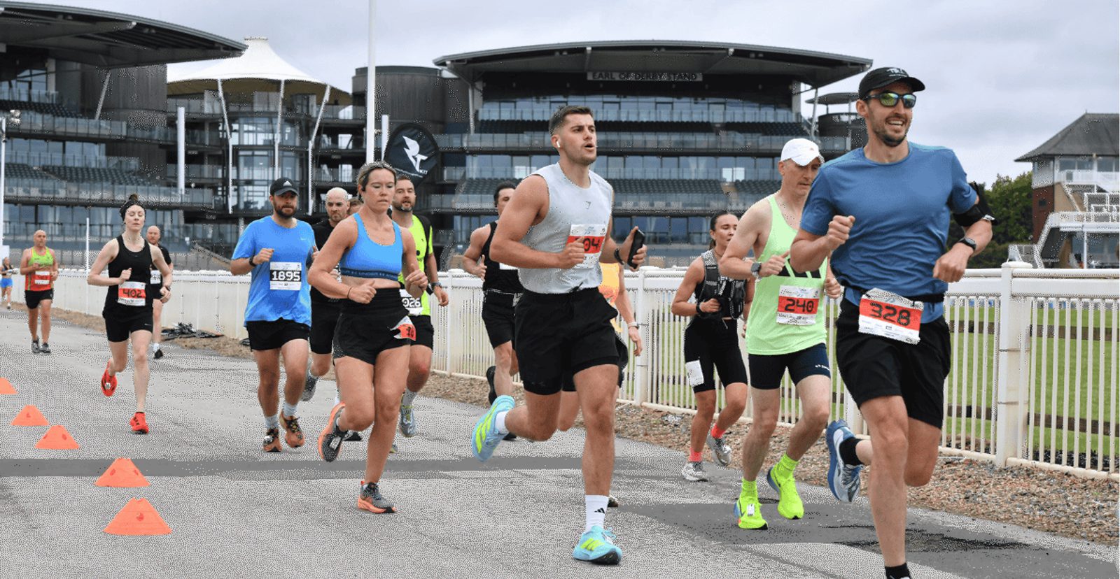 A group of runners wearing race bibs participate in a road race, running along a track with orange cones and modern buildings visible in the background. The sky is cloudy.