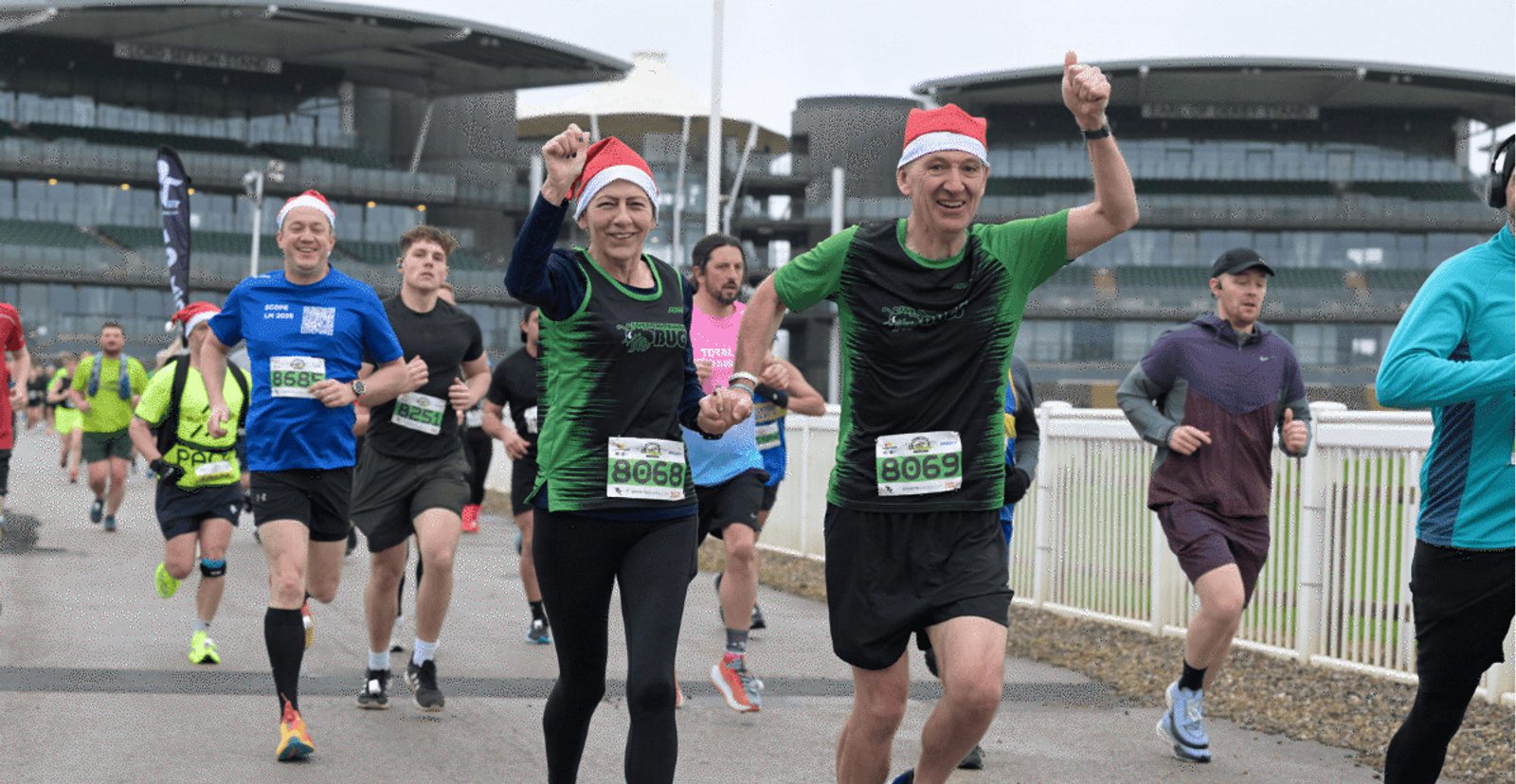 A group of runners, some wearing Santa hats, participate in a race. Two runners in green shirts smile and raise their arms in celebration. Large modern buildings are visible in the background.