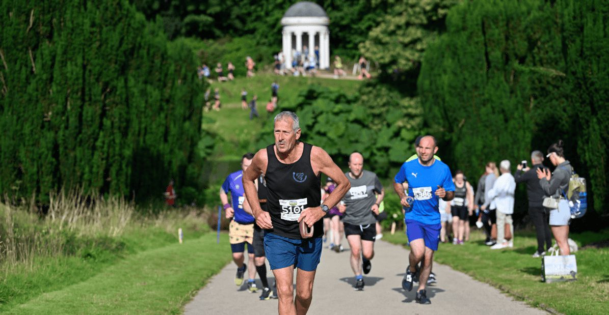 Runners participate in a race on a path through a green park, with a domed pavilion on a hill in the background and spectators standing beside the route.
