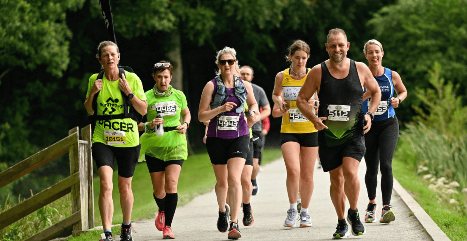 A group of runners, both men and women, jog on a paved path in a park. They wear race bibs and athletic gear, with trees and greenery in the background. Everyone looks focused and determined.