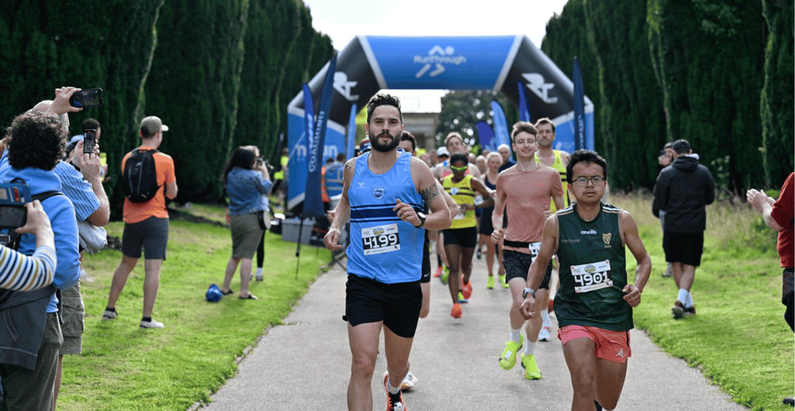 Runners in numbered bibs participate in a race, passing under a blue inflatable archway, while spectators stand on either side of the path, clapping and taking photos.