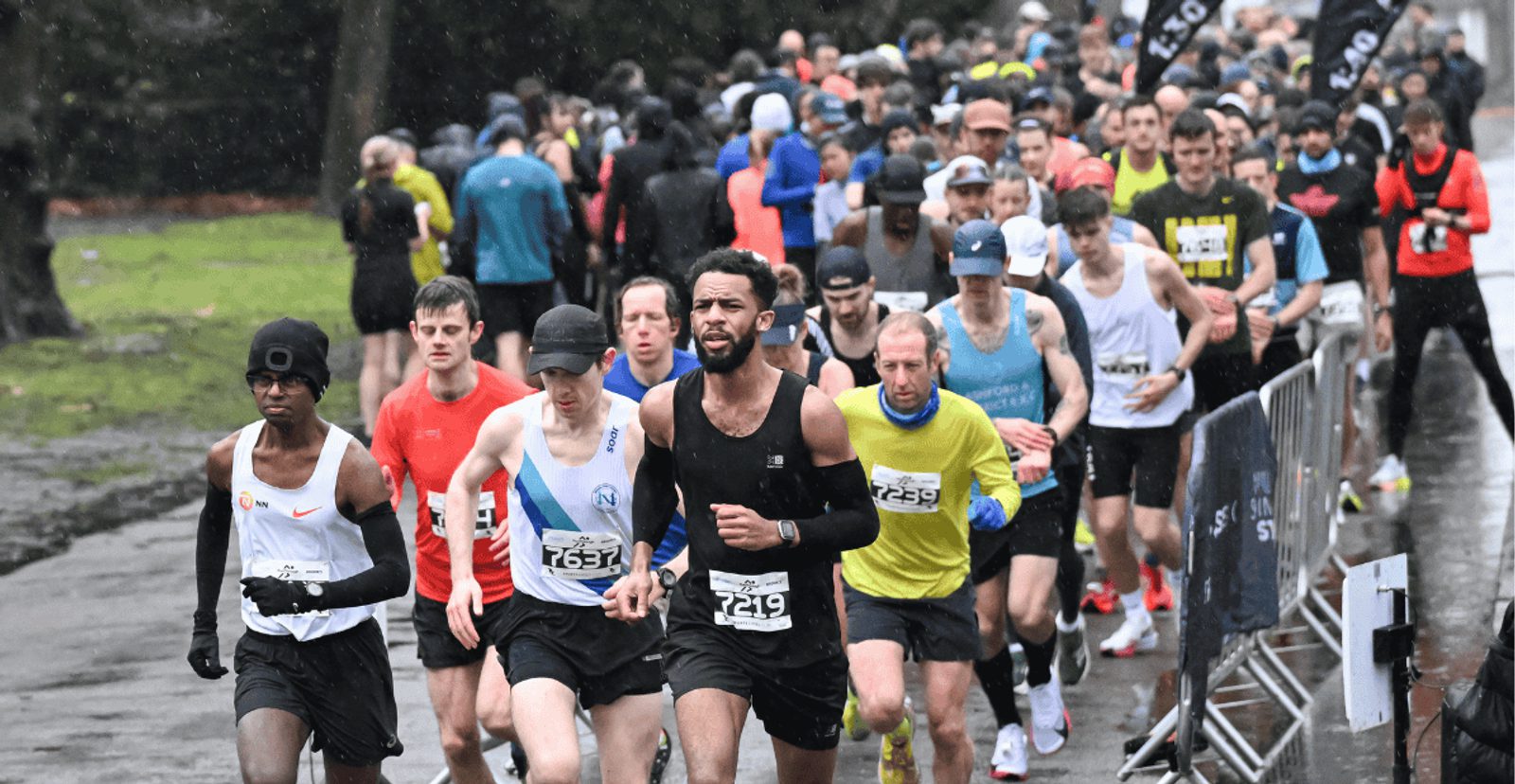 A large group of runners in athletic gear participate in a race on a wet road, with some wearing race bibs and others in rain jackets. Spectators and trees are visible in the background.