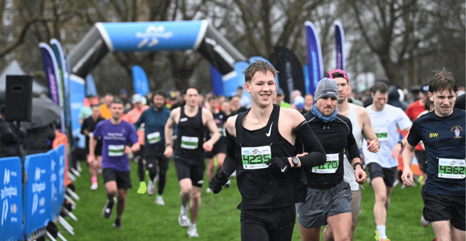 A group of runners, wearing race bibs, start a race on a grassy field under an inflatable archway on a cloudy day. The participants look focused and energetic.