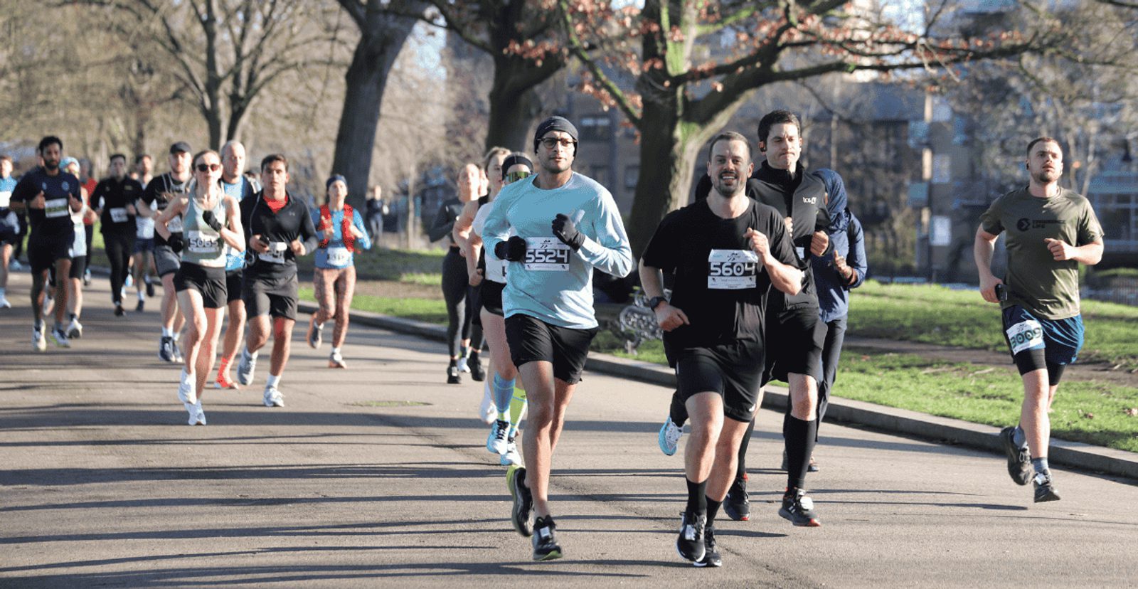 A group of runners, each wearing numbered bibs, runs along a paved path in a park on a sunny day. Trees line the route, and the participants appear focused and energetic.