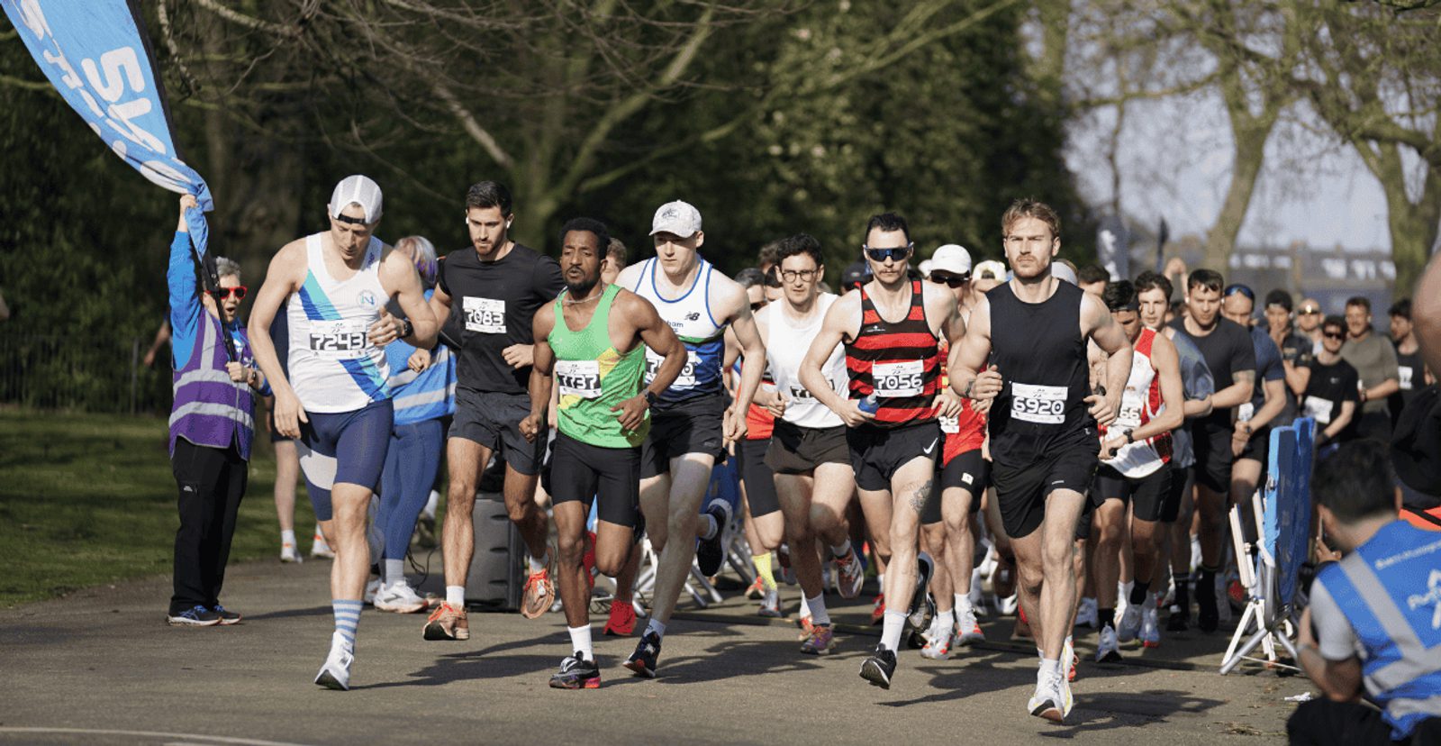 A group of runners in athletic gear start a race outdoors on a sunny day, with trees and spectators visible in the background.