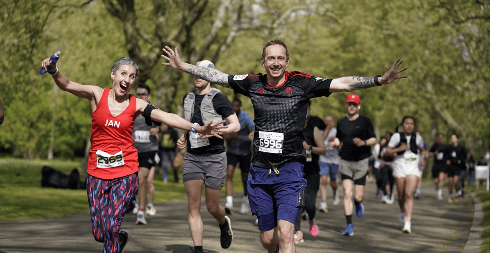 Two smiling runners with outstretched arms, one in a red tank top and the other in black, lead a group of people running outdoors on a tree-lined path during a race.