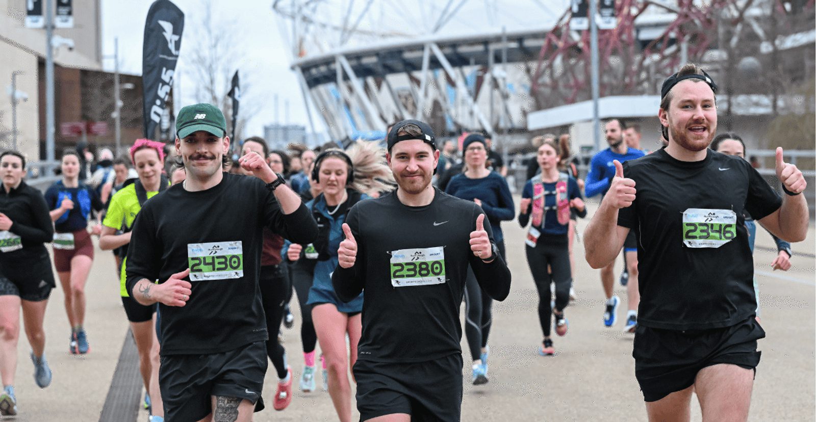 A group of runners, many smiling and giving thumbs up, participate in a race. Most wear black athletic gear and numbered bibs. A stadium and other runners are visible in the background.