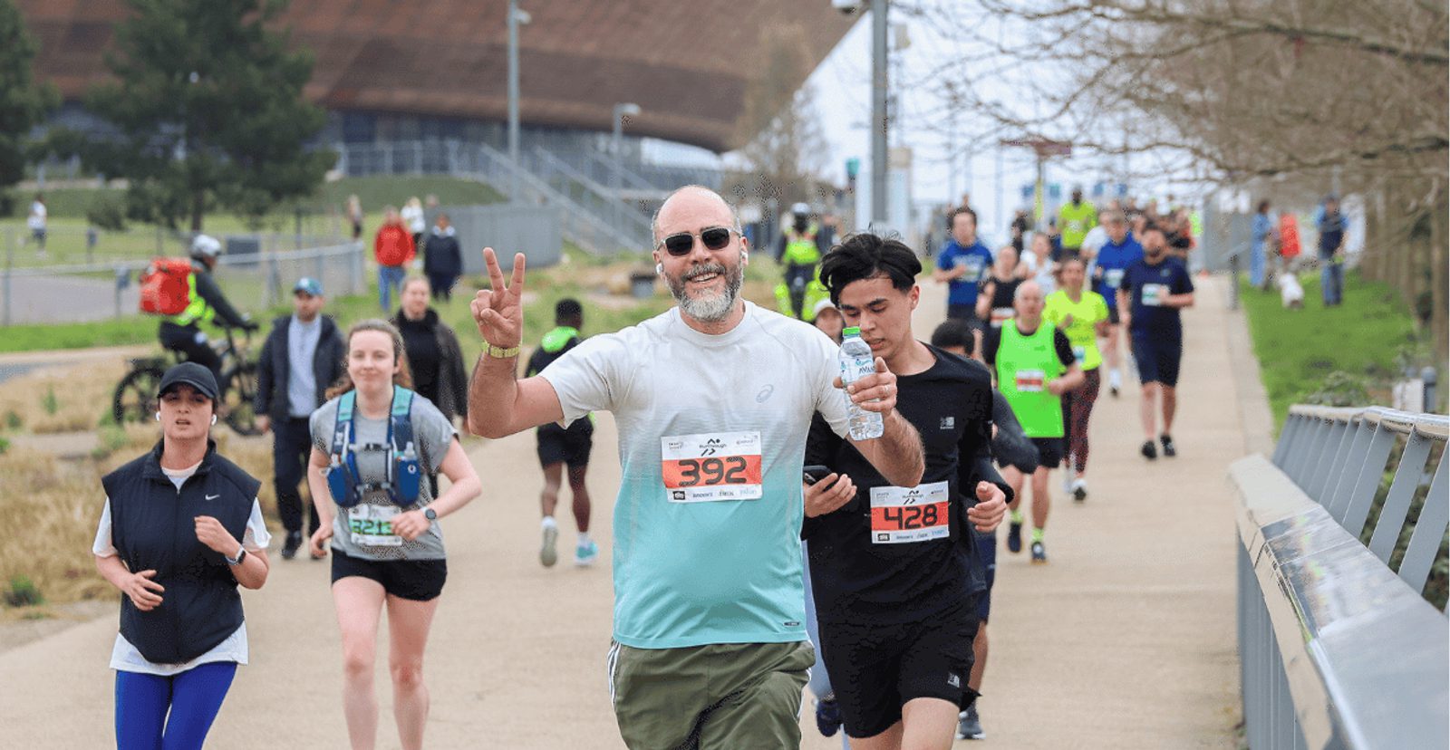 A group of runners participate in a race on a paved path. A smiling man in front, wearing sunglasses and race bib 392, holds up a peace sign and a water bottle. Other runners follow behind him.