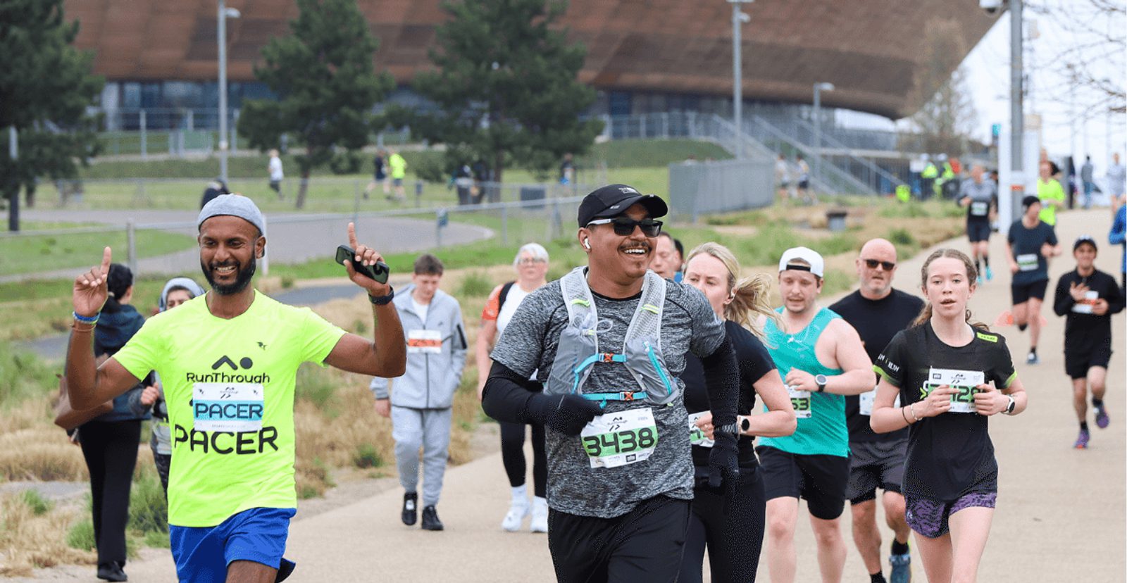 A group of runners participating in a race outdoors. A smiling pacer in a neon shirt leads, showing two fingers up, while other runners follow behind, some smiling and others focused. A large building is visible in the background.