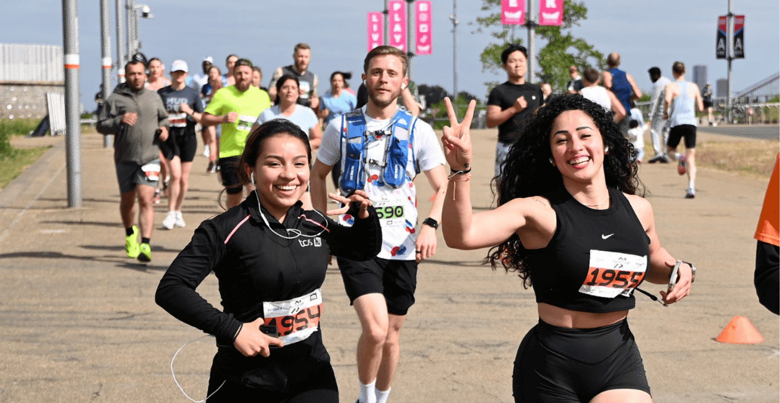 Two women smiling and running in a race, both wearing race bibs. One woman flashes a peace sign. Other runners follow behind on a sunny day.