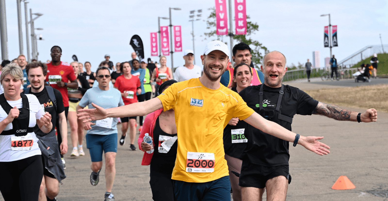 A smiling man in a yellow shirt and white cap, with arms wide open, leads a group of runners during a race on a paved path. Other runners follow behind him, some also smiling and raising their arms.
