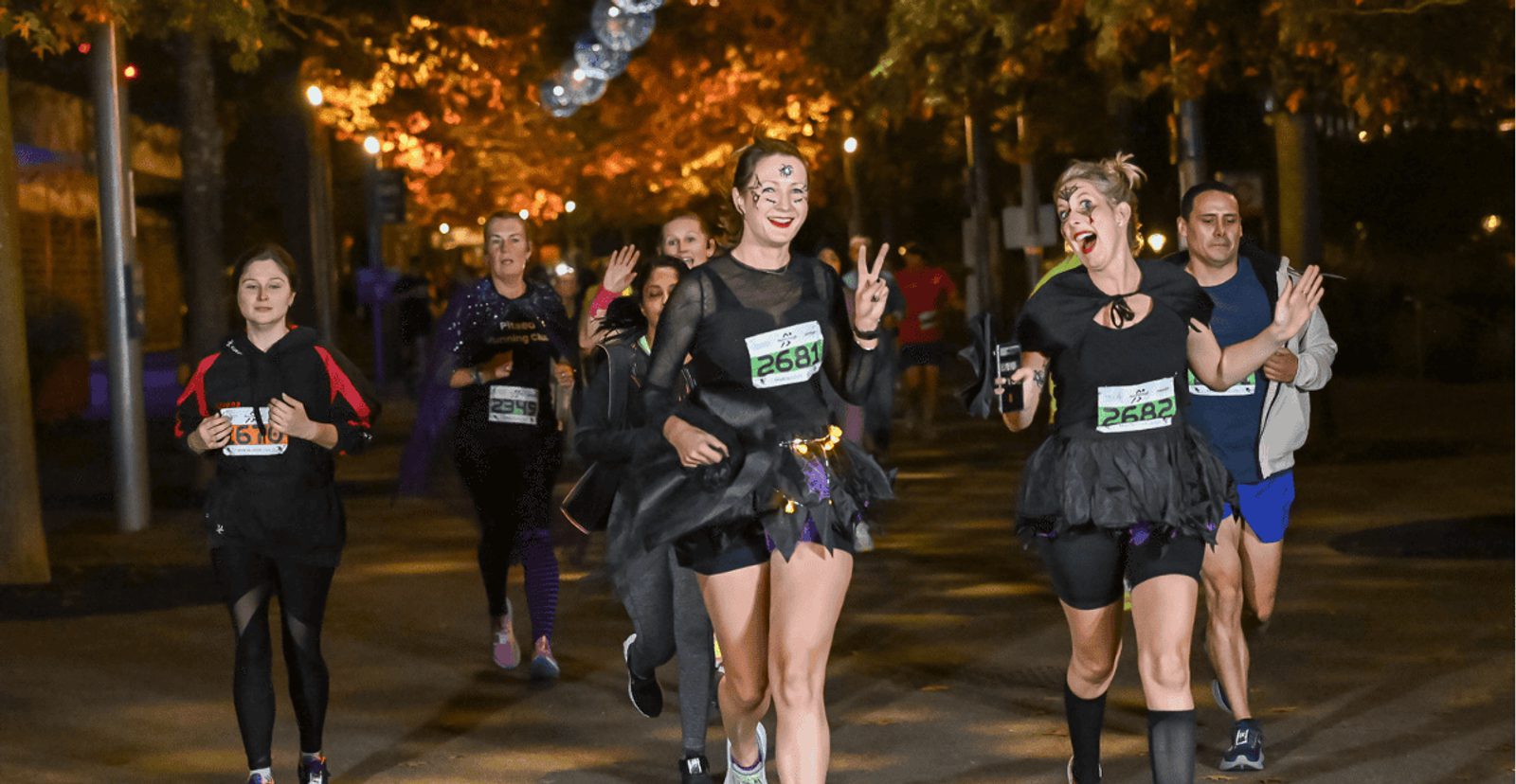 A group of people in costumes run at night during a race. Two women in front wear black tutus and smile, one flashing a peace sign. Others follow behind under streetlights and autumn trees. Race bibs are visible on their outfits.