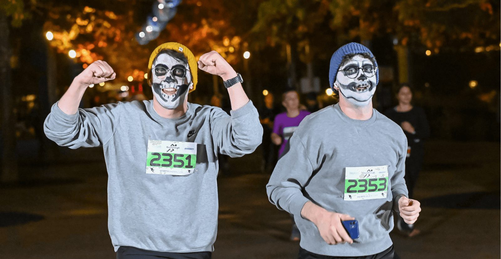Two runners wearing skeleton face paint, gray sweatshirts, and numbered race bibs (2351 and 2353) smile and pose during a night race, with blurred participants and autumn trees in the background.