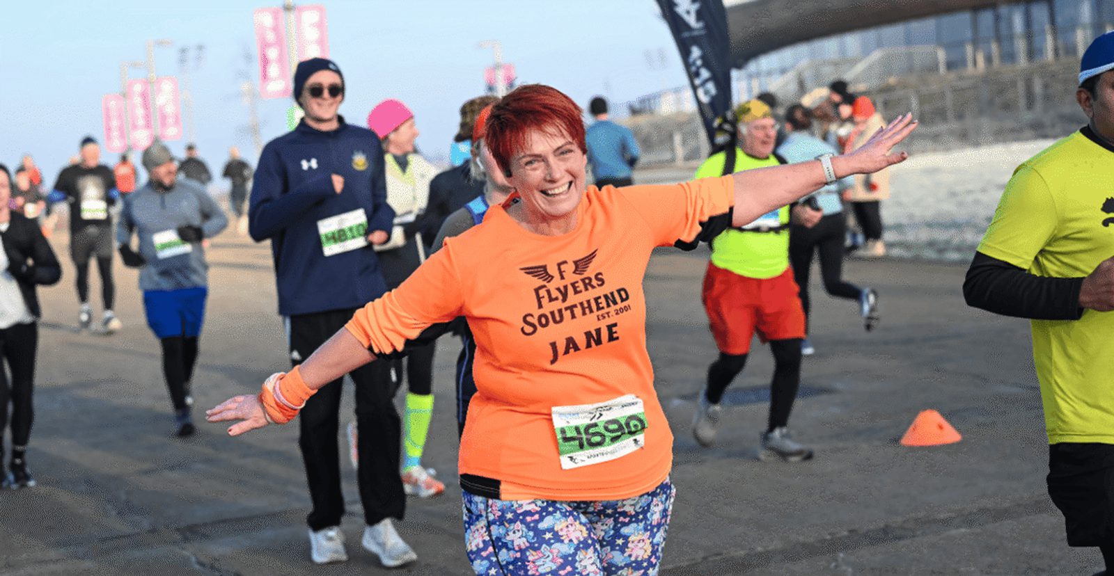 A woman with red hair and a big smile, wearing an orange "Flyers Southend Jane" shirt and patterned leggings, runs with her arms outstretched among other runners on a paved course.