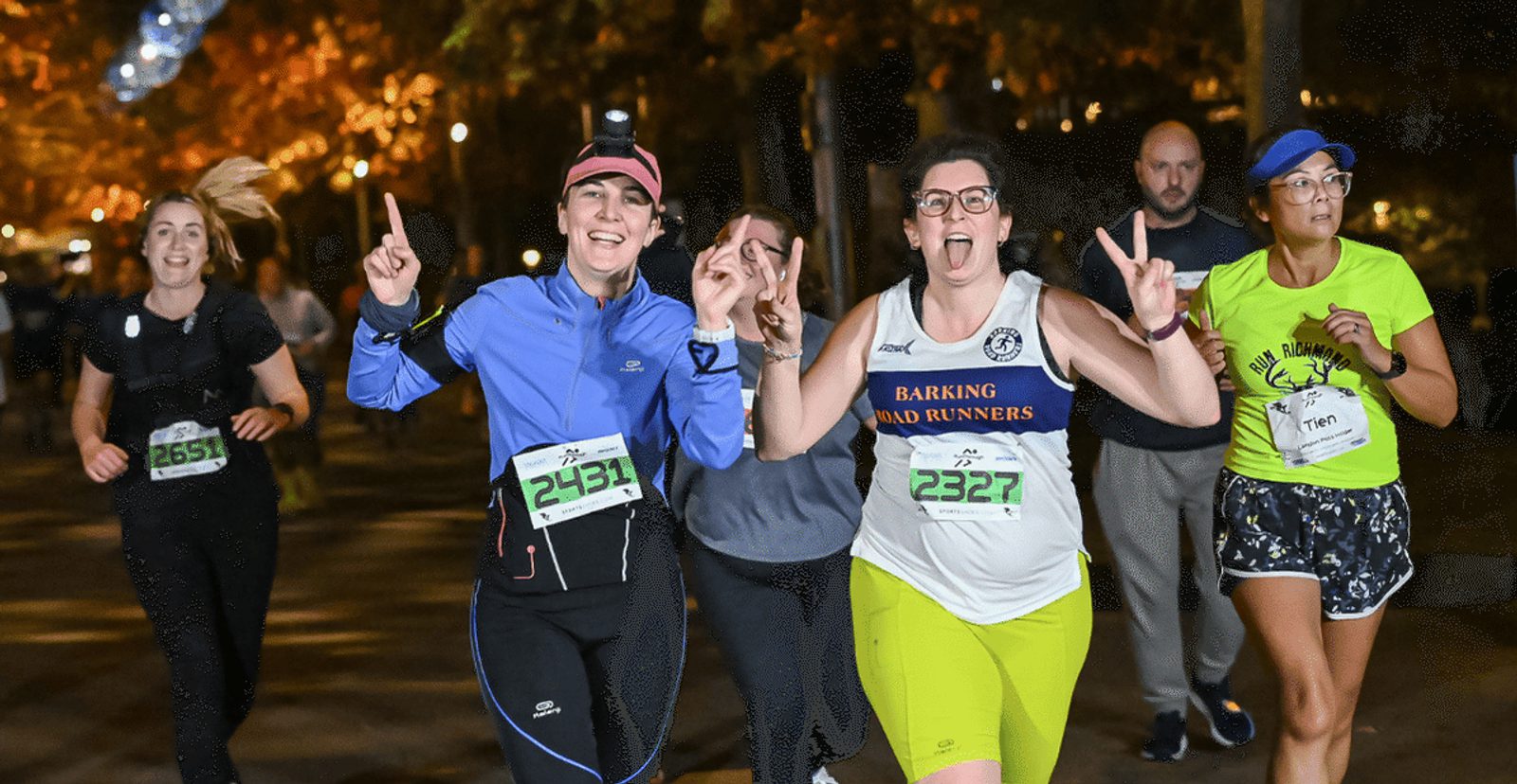 A group of runners, some smiling and making peace signs, participate in a nighttime race under streetlights, wearing numbered bibs and athletic gear. Trees with autumn leaves line the path.