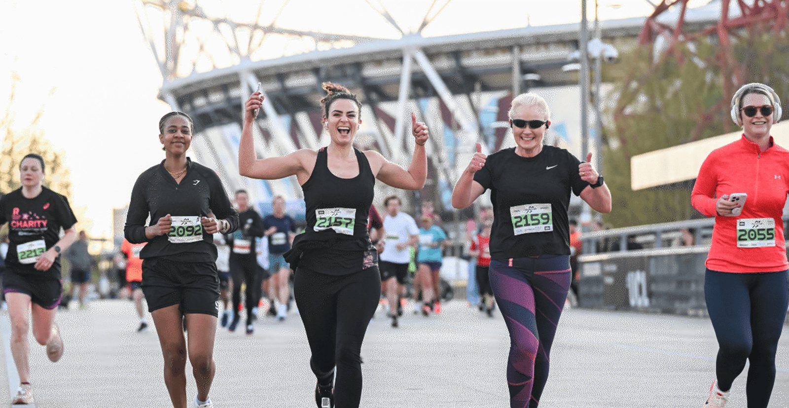 A group of runners, mostly women, smile and give thumbs up as they participate in a race outdoors, with a stadium structure visible in the background. They wear race bibs and athletic clothing.