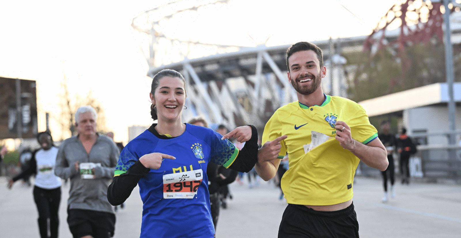 Two runners smile and point at each other's Brazilian soccer jerseys during a race. Other participants run in the background near a stadium structure on a sunny day.