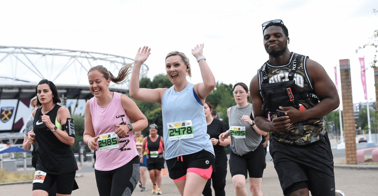 A group of runners, both men and women, participate in a race outdoors. One woman in a blue shirt with race bib 2478 smiles with her arms raised, while others run or smile nearby. A stadium is visible in the background.