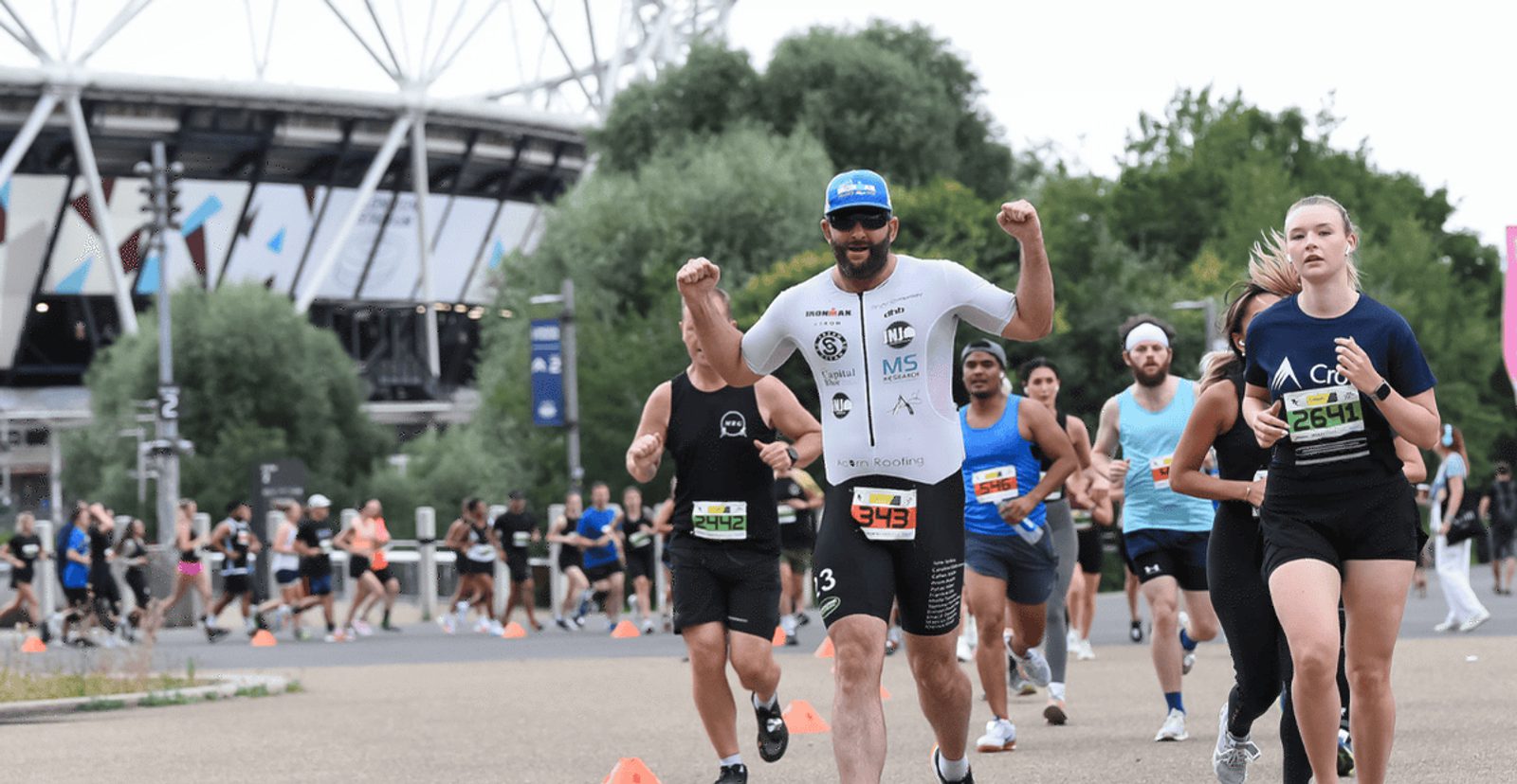 A group of runners participates in a race near a stadium. One man in front, wearing sunglasses and a blue cap, raises his arms in celebration as others run behind him. Trees and spectators are visible in the background.