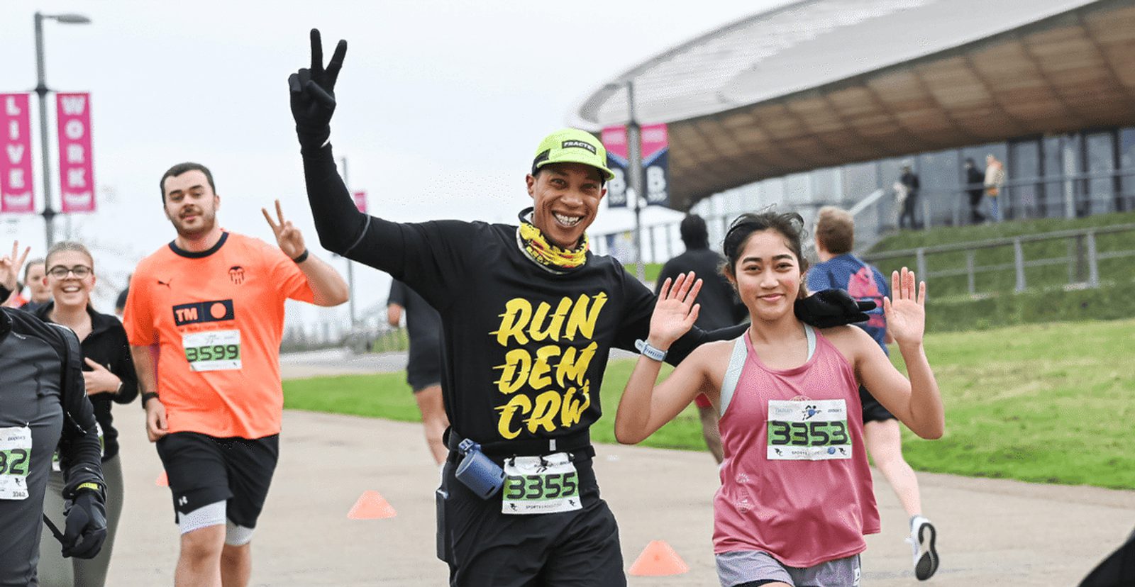 A smiling man and woman wearing race bibs wave as they run outdoors with other participants. The man wears a "Run Dem Crew" shirt and a neon cap; the group appears happy and energetic near a modern building.