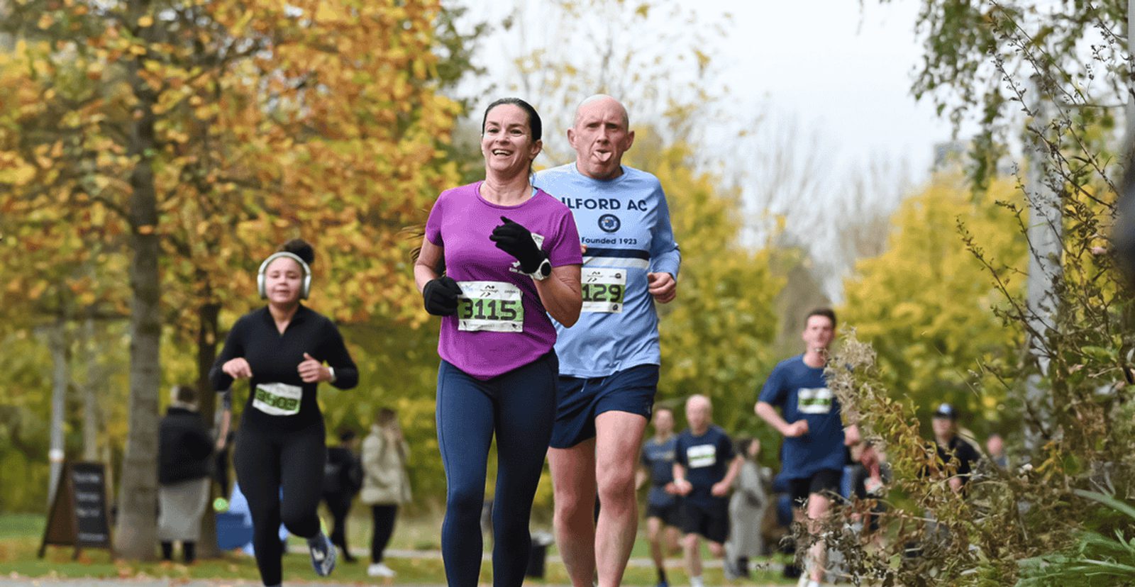 A woman and a man, both wearing race bibs, run outdoors on a tree-lined path with other runners in the background. The woman is smiling, and the trees display green and yellow autumn foliage.