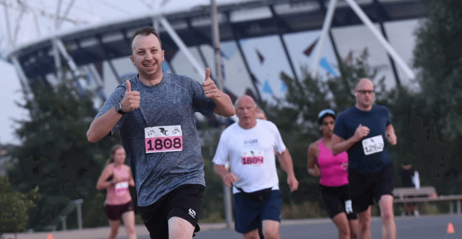 A man in a gray shirt and black shorts runs towards the camera, smiling and giving thumbs up, with other runners behind him. All have numbered race bibs. A stadium is visible in the background.