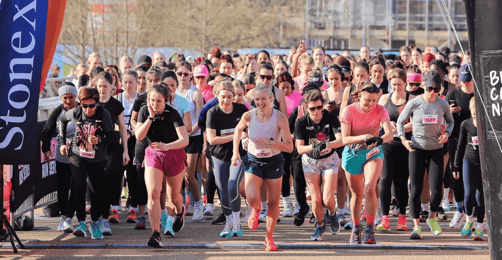 A large group of women in athletic wear start running at the beginning of a race, with excited expressions and sunlight shining on them. Spectators and banners are visible in the background.